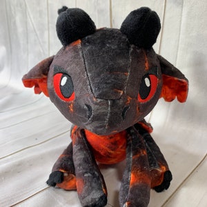 Puede incluir: Un peque&ntilde;o drag&oacute;n de peluche negro y naranja con ojos rojos. El drag&oacute;n est&aacute; sentado sobre una superficie de madera blanca.