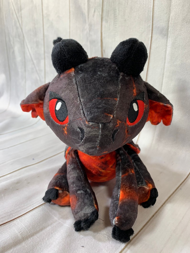 Puede incluir: Un peque&ntilde;o drag&oacute;n de peluche negro y naranja con ojos rojos. El drag&oacute;n est&aacute; sentado sobre una superficie de madera blanca.