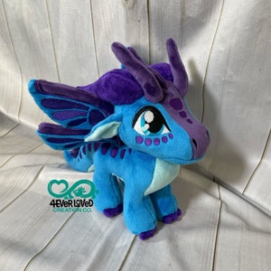 K&ouml;nnte beinhalten: Ein Pl&uuml;schdrache in Blau und Lila mit gro&szlig;en Fl&uuml;geln, blauen Augen und einem lila-blauen Muster auf dem K&ouml;rper. Der Drache steht auf einer Holzoberfl&auml;che. 4ever Loved Creation Co. ist auf der Oberfl&auml;che aufgedruckt.