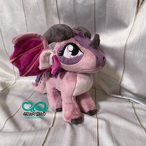 K&ouml;nnte beinhalten: Ein rosa Pl&uuml;schdrache mit violetten Akzenten und einem rosa-wei&szlig; gestreiften Fl&uuml;gel. Der Drache hat ein graues Horn und gro&szlig;e violette Augen. Der Drache steht auf einer wei&szlig;en Holzoberfl&auml;che.