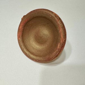 Può includere: Un piccolo vaso rotondo in terracotta con una glassa marrone. Il vaso è vuoto e ha una texture leggermente ruvida.