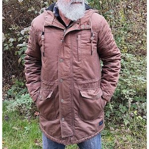 Op de afbeelding: Een bruine capuchonjas met een rits en knoopsluiting. De jas heeft een capuchon met trekkoord, twee voorzakken en een klepzak op de linkermouw.