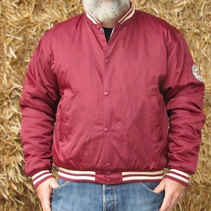 Könnte beinhalten: Eine weinrote Satin-Bomberjacke mit weißen Rippbündchen an Kragen, Ärmelbündchen und Bund. Die Jacke hat einen Knopfverschluss und einen kleinen Aufnäher am rechten Ärmel.