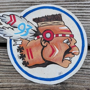 Op de afbeelding: Een handgeschilderde ronde sticker met het gezicht van een indiaan met rode, witte en zwarte verf. De man heeft een blauwe verenhoed en een blauwe pijl achter zich. De sticker is omgeven door een blauwe cirkel.