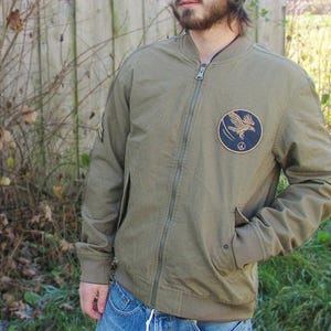 Könnte beinhalten: Eine grüne Canvas-Bomberjacke mit Reißverschluss und einem Patch auf der linken Seite mit einem fliegenden Vogel und einem Peace-Zeichen.