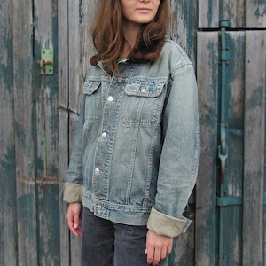Könnte beinhalten: Eine hellblaue Jeansjacke mit einem braunen Kunstfellkragen. Die Jacke hat einen Knopfverschluss und wird von einer Person getragen, die eine schwarze Newsboy-Mütze und eine Sonnenbrille trägt.