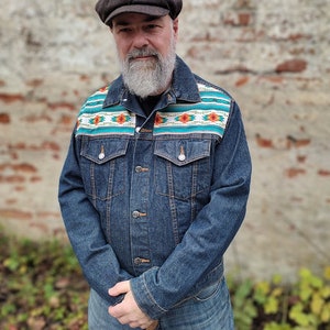 Può includere: Una giacca di jeans blu con un motivo geometrico colorato sul retro. La giacca ha una chiusura con bottoni e due tasche sul davanti. La persona che indossa la giacca indossa anche un cappello da newsboy marrone.