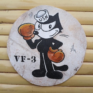 Op de afbeelding: Een rond, handgeschilderd kunstwerk met de stripfiguur Felix the Cat, met een matrozenhoed en bokshandschoenen. Het kunstwerk bevat de tekst "VF-3" en "USN" op de hoed, met een verweerde, verouderde uitstraling.