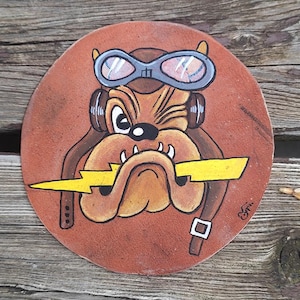 Könnte beinhalten: Ein runder, brauner Untersetzer mit einem Cartoon-Bulldoggen-Design. Der Bulldogge trägt eine Fliegerbrille und ein Headset und hält einen gelben Blitz. Der Untersetzer ist auf einer Holzoberfläche bemalt.