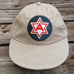 Peut inclure: Une casquette de baseball beige avec un écusson brodé noir et rouge représentant une étoile et la lettre "A".