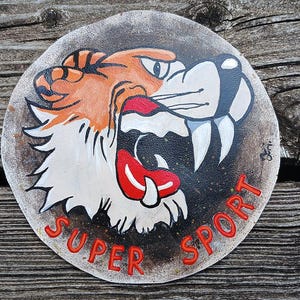 Può includere: Emblema rotondo dipinto a mano raffigurante la testa di una tigre ruggente con dettagli arancioni, bianchi e neri. Le parole "SUPER SPORT" sono rosse, bordate di nero. L'emblema è su uno sfondo circolare usurato.