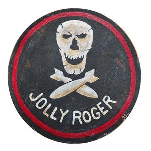Op de afbeelding: Ronde, zwarte Jolly Roger-embleem met een witte schedel en gekruiste beenderen, en twee gekruiste bommen. Het embleem heeft een rode rand en de woorden "JOLLY ROGER" in het wit.