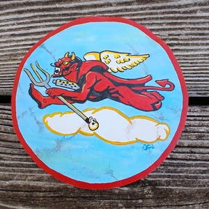 Peut inclure: Œuvre d'art ronde peinte à la main représentant un diable rouge avec un trident, des ailes et une queue, courant dans un ciel bleu avec un nuage blanc. L'œuvre a une bordure rouge et est posée sur un fond en bois.