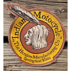 Puede incluir: Una pegatina redonda amarilla y roja con el texto "The Indian Motorcycle Co. The Hendee Manufacturing Springfield Mass." y un dibujo de la cabeza de un hombre nativo americano con un tocado de plumas.