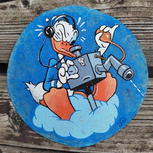 Peut inclure: Une pancarte ronde en bois peinte à la main représentant un canard de dessin animé portant un casque et tenant un fusil. Le canard est assis sur un nuage sur un fond bleu.