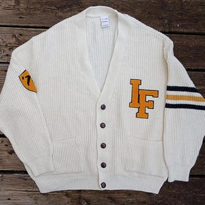 Op de afbeelding: Crèmekleurig vest met knopen, een V-hals en twee voorzakken. Voorzien van een geel en oranje "LF"-patch op de borst en een "'7"-patch op de linkermouw. De rechter mouw heeft gele en marineblauwe strepen.