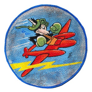 Op de afbeelding: Een vintage leren patch met een cartoon wolf-piloot die een rood vliegtuig bestuurt. De wolf draagt een groene helm en een bril. Het vliegtuig heeft gele bliksemschichten erachter. De patch is blauw met een witte achtergrond.
