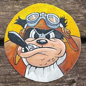 Op de afbeelding: Een ronde, geschilderde leren patch met een cartoonhond die een vliegeniersbril en een leren helm draagt. De hond heeft een sigaar in zijn bek en een serieuze uitdrukking. De achtergrond is geel.