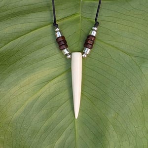 May include: A necklace featuring a long, smooth, off-white tapered pendant suspended from a black cord. The design includes two sets of dark brown cylindrical beads and metallic silver spacers positioned symmetrically on each side of the centre piece.