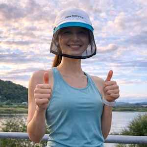 May include: A white New Balance visor with a blue brim and a black mesh face shield. The visor is worn with a light blue tank top. The person is giving a thumbs up.