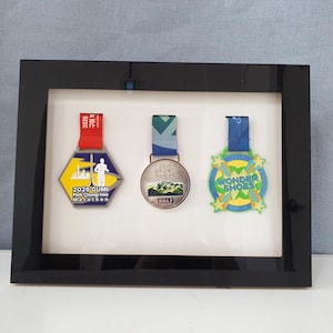 May include: A black rectangular display frame containing three medals with ribbons. The left medal is hexagonal with a red ribbon, the center is circular with a blue and green ribbon, and the right is star-shaped with a blue ribbon and text reading WONDER SHOES.