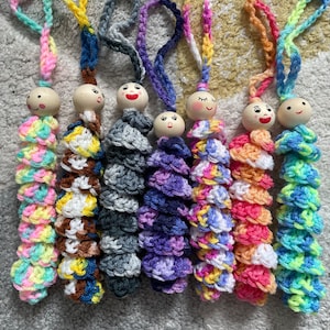May include: Seven colourful crocheted doll keychains with wooden heads and painted faces. Each keychain features a different colour combination and pattern.