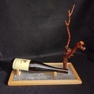 May include: A brown wine bottle with a yellow label rests on a wooden tray with a clear surface. A decorative wooden branch stands upright next to the bottle. The background is dark.