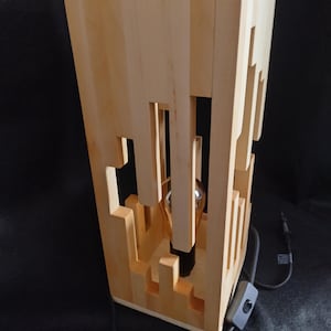 May include: A wooden table lamp with a geometric design. The lamp has a light-colored wood frame with cut-out shapes, and a visible lightbulb. The lamp includes a black power cord and a switch. The lamp is approximately 12 inches tall.