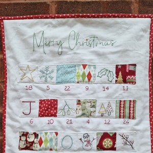 Peut inclure: Calendrier de l'Avent de Noël fait à la main, avec un fond blanc et une bordure rouge. Le calendrier présente des motifs brodés et les mots "Merry Christmas". Chaque jour a un design et un numéro uniques.