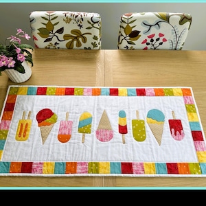 May include: A rectangular quilted placemat featuring a white center panel decorated with a row of colorful ice cream cones and popsicles in shades of red, yellow, green, blue, and pink, framed by a border of multi-colored fabric squares.