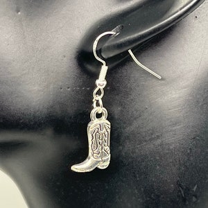 May include: Silver dangle earrings featuring a small boot charm. The boot is detailed with a textured design.