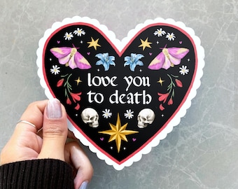 May include: Heart-shaped sticker with a black background and a scalloped edge. The sticker features the text "love you to death", two skulls, moths, flowers, stars, and a gold starburst.