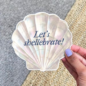 May include: A seashell-shaped sticker with iridescent colors and the text "Let's shellebrate!" in navy blue. The sticker is held by a hand with a light purple painted fingernail.