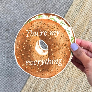 Può includere: Un adesivo rotondo raffigurante un bagel con formaggio cremoso ed erba cipollina. Il testo "You're my everything" è scritto in bianco. L'adesivo è tenuto da una mano con un'unghia dipinta di viola chiaro.