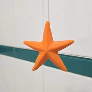 May include: A vibrant orange starfish suspended against a backdrop of white and teal tiles. The five-pointed star is hanging by a thin string, creating a floating effect. The starfish is a solid colour.