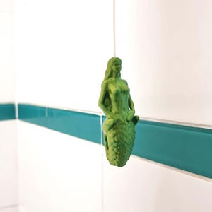 May include: A green mermaid figurine with a detailed design, standing on a tiled surface with teal grout.