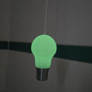 May include: A green light bulb pendant with a silver base suspended from a thin white cord. The bulb emits a soft green light. The background is a neutral grey with a dark green horizontal stripe.