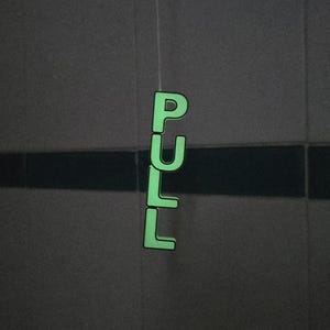 May include: A luminous green sign spelling "PULL" hangs against a dark tiled wall. The capital letters are arranged vertically and seem to be lit up, creating a striking contrast.