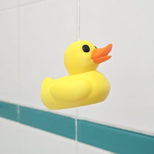 May include: A yellow rubber duck with black eyes and an orange beak hangs from a string. The duck is suspended against a white tiled background with a teal accent stripe.
