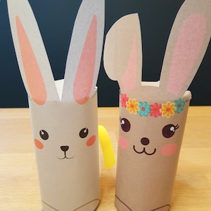 以下が含まれることがあります： 段ボールチューブで作られた2つの手作りのウサギクラフト。1匹のウサギは黄色のリボン、もう1匹は花のヘッドバンドをしています。どちらのウサギも、ピンク色の内耳、バラ色の頬、そしてかわいい顔の特徴を持っています。