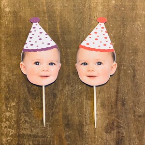 K&ouml;nnte beinhalten: Zwei Cupcake-Topper mit einem Babygesicht, das einen Partyhut tr&auml;gt. Die H&uuml;te sind wei&szlig; mit roten und lila Punkten. Die Topper sind auf Holzst&auml;bchen.