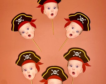 May include: A collection of baby face cutouts on wooden sticks, each wearing a pirate hat with a skull and crossbones emblem. The hats are black with yellow trim and red bandanas. The background is a solid, muted orange.