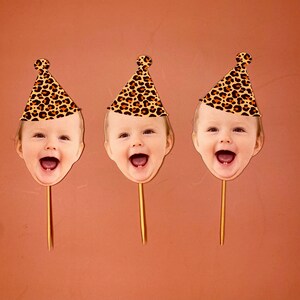 May include: Three cake toppers featuring a child's face with a leopard print party hat. Each topper is on a wooden stick.