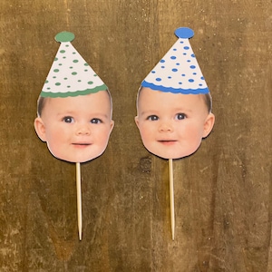 K&ouml;nnte beinhalten: Zwei Tortendekorationen mit einem Babygesicht und einer Partym&uuml;tze. Eine M&uuml;tze ist gr&uuml;n mit wei&szlig;en Punkten und die andere ist blau mit wei&szlig;en Punkten.