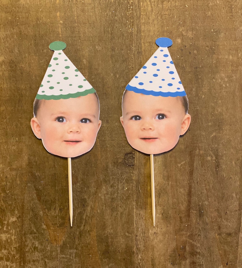 K&ouml;nnte beinhalten: Zwei Tortendekorationen mit einem Babygesicht und einer Partym&uuml;tze. Eine M&uuml;tze ist gr&uuml;n mit wei&szlig;en Punkten und die andere ist blau mit wei&szlig;en Punkten.