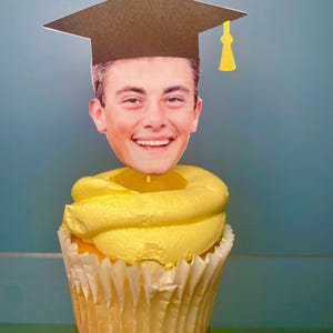 May include: A yellow cupcake with white frosting topped with a graduation cap and a photo of a person's face.