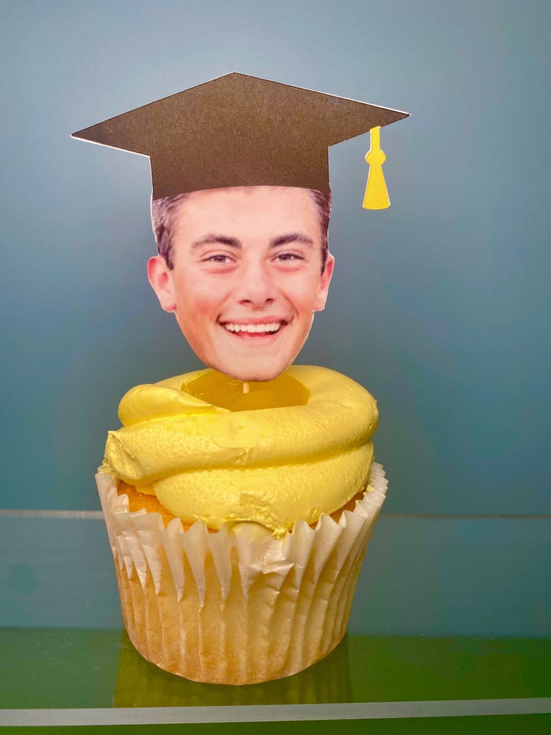 May include: A yellow cupcake with white frosting topped with a graduation cap and a photo of a person's face.
