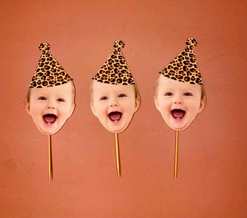 May include: Three cake toppers featuring a child's face with a leopard print party hat. Each topper is on a wooden stick.
