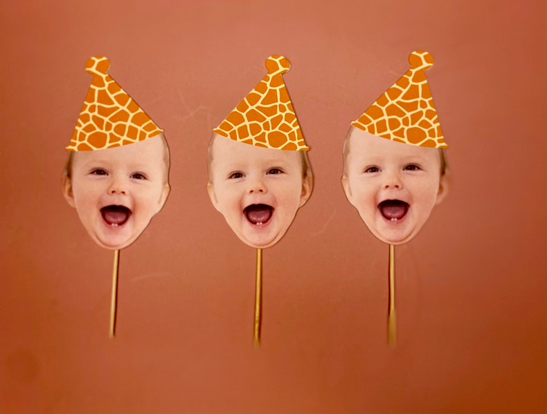 May include: Three orange and brown giraffe print party hats with a baby's face cut out in the front. The hats are on sticks.
