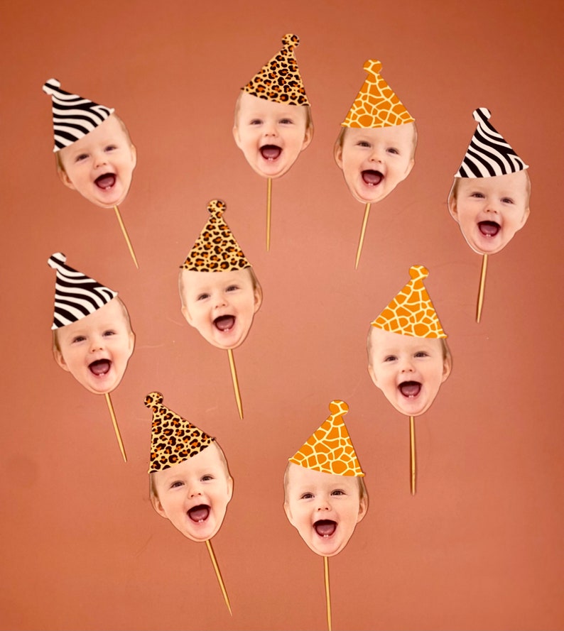 May include: Nine cupcake toppers featuring different animal print party hats with a baby's face in each hat. The hats are in various animal prints, including zebra, giraffe, and leopard.